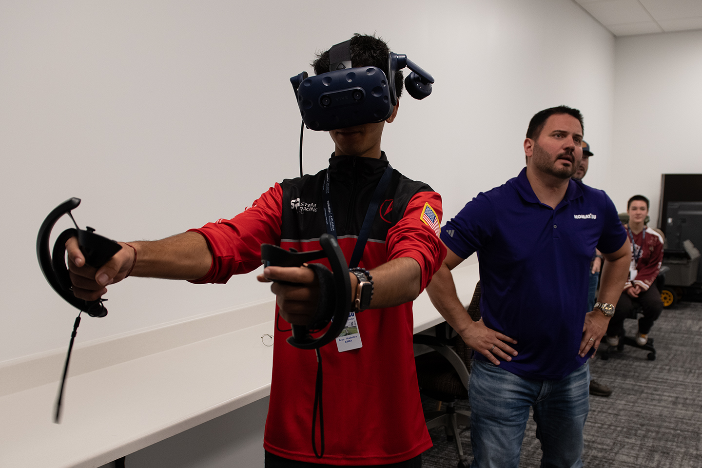 Participant wearing a virtual reality headset and handheld controllers takes part in an immersive technology demonstration while observers watch in a training room.