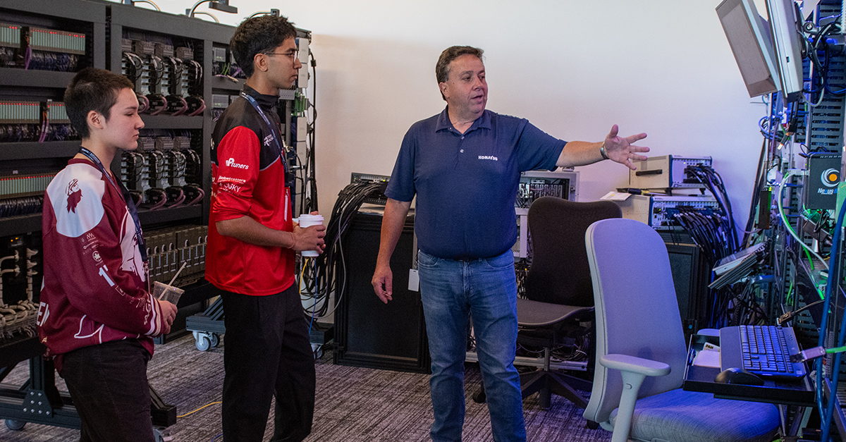 Two students tour a technology lab as a Komatsu employee explains computer systems and network equipment housed in server racks.