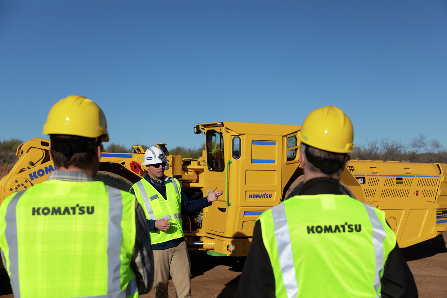 Workers wearing high-visibility Komatsu safety vests and hard hats participate in an outdoor equipment briefing near large yellow Komatsu mining machines.