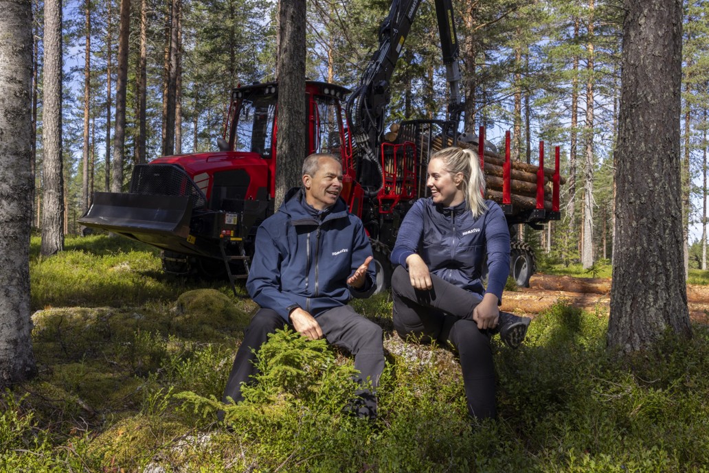 Two people sitting on a forest floor beside a red forestry machine loaded with cut logs, surrounded by tall trees in a wooded area.
