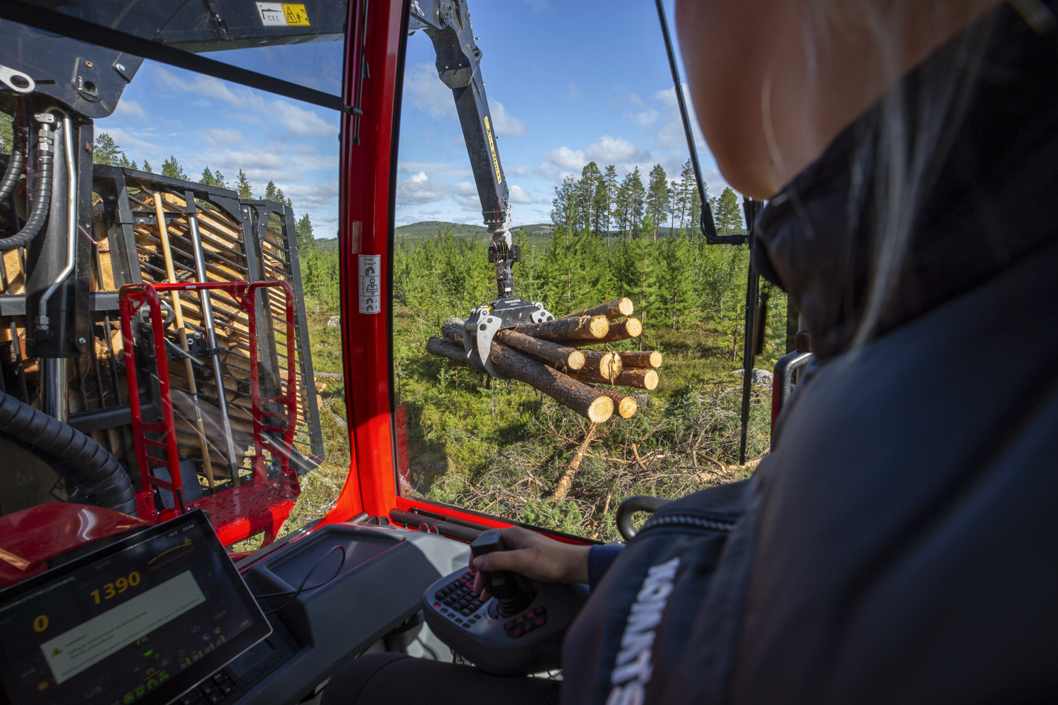 View from inside the cab of a forestry machine as an operator uses joystick controls to move a hydraulic crane carrying a bundle of logs.