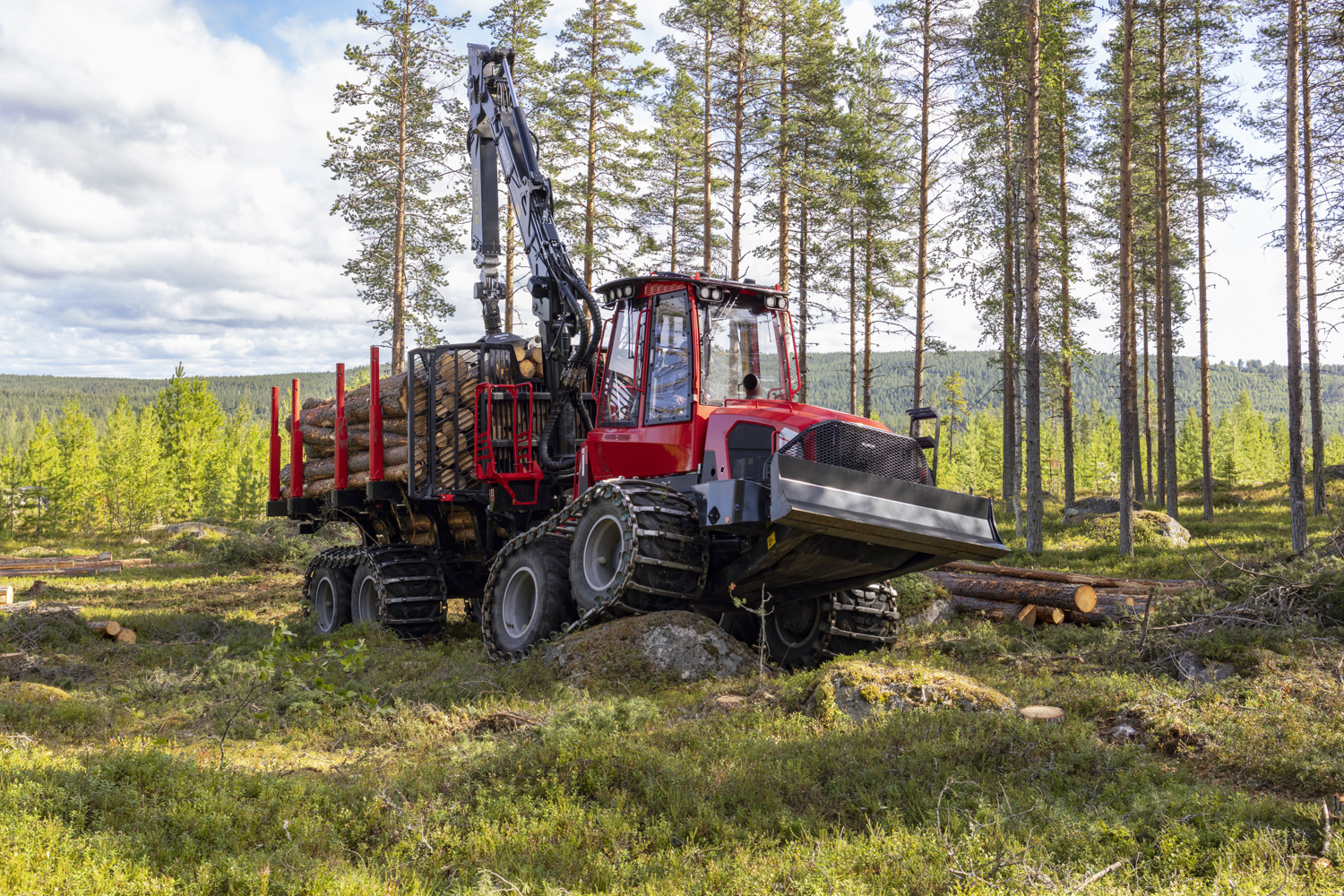 Red forestry machine loaded with cut logs stands in a partially harvested forest clearing.