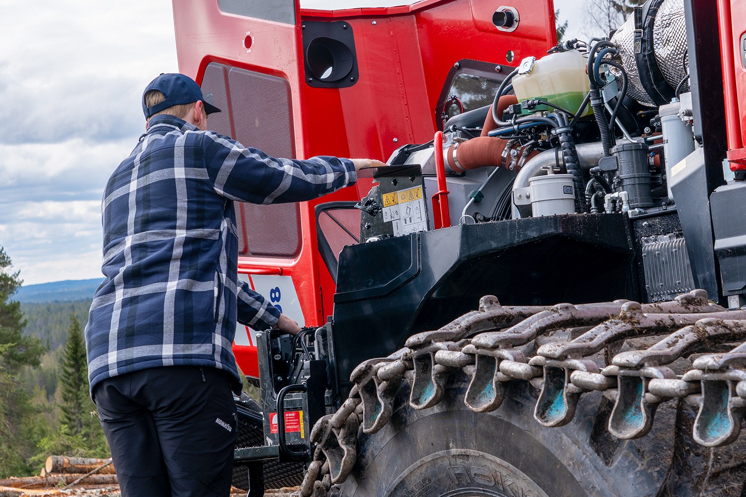 A worker performs maintenance on a red forestry machine with an open engine compartment.