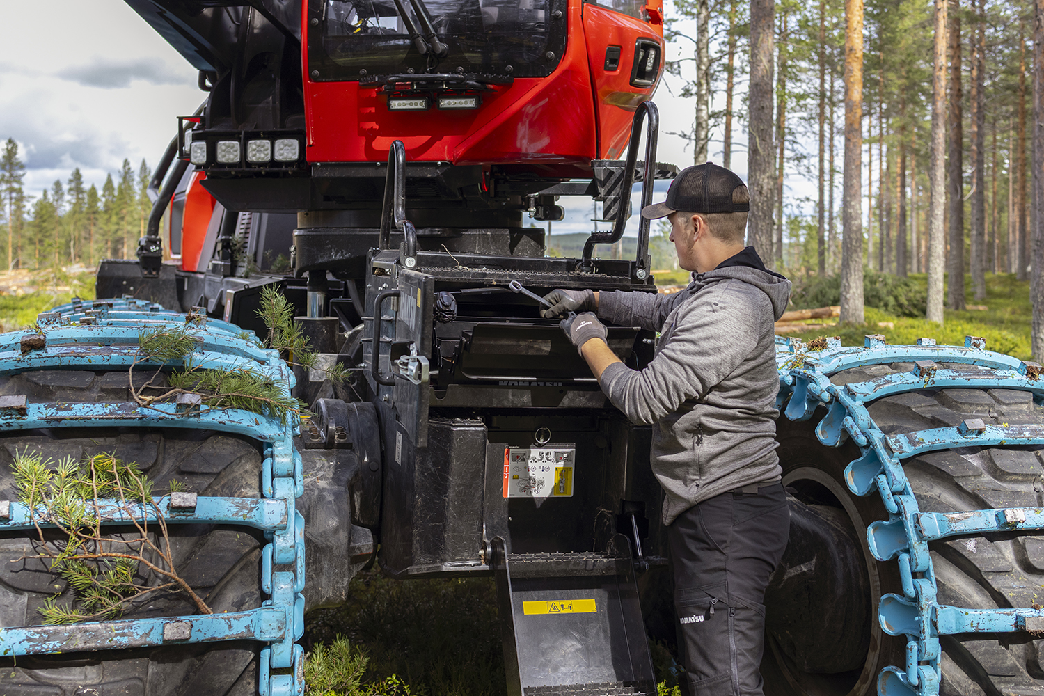 A person performs maintenance on a red forestry machine in a forest clearing.