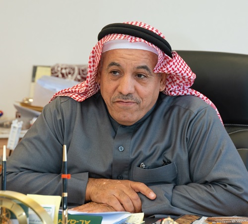 علي ح. العيوني يرتدي الزي التقليدي للشرق الأوسط (الكوفية والثوب) جالسًا على مكتب في مكتبه محاطًا بالوثائق والعناصر الديكورية