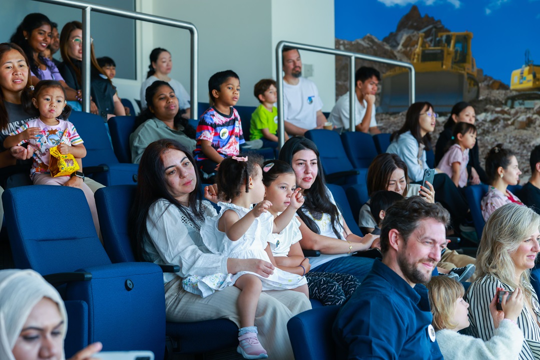 جمهور جالس على كراسي زرقاء في قاعة، يشاهد عرضًا تقديميًا؛ يضم مزيجًا من البالغين والأطفال