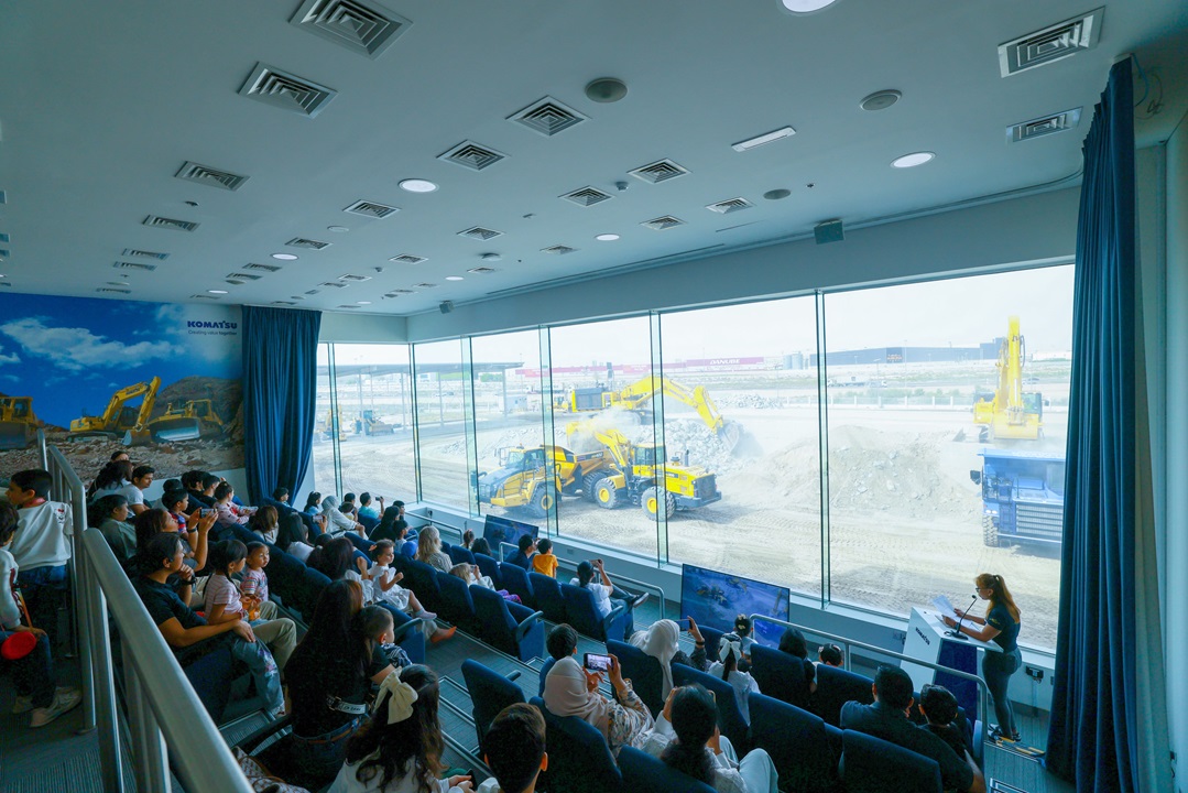 جمهور في غرفة ذات جدران زجاجية يشاهد عرضًا حيًا لأعمال البناء؛ آلات Komatsu الصفراء تعمل في الخارج
