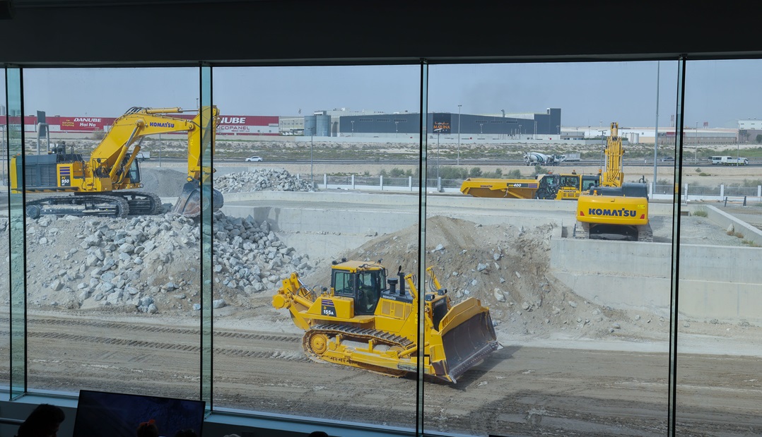 ثلاث آليات Komatsu صفراء — آليتا حفر وجرار — تعمل في موقع بناء خلف حاجز زجاجي، حيث تقوم بنقل التربة والصخور.