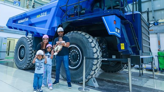 يقف خمسة أشخاص، بينهم ثلاثة أطفال، أمام شاحنة تفريغ زرقاء ضخمة من طراز Komatsu داخل مرفق صناعي