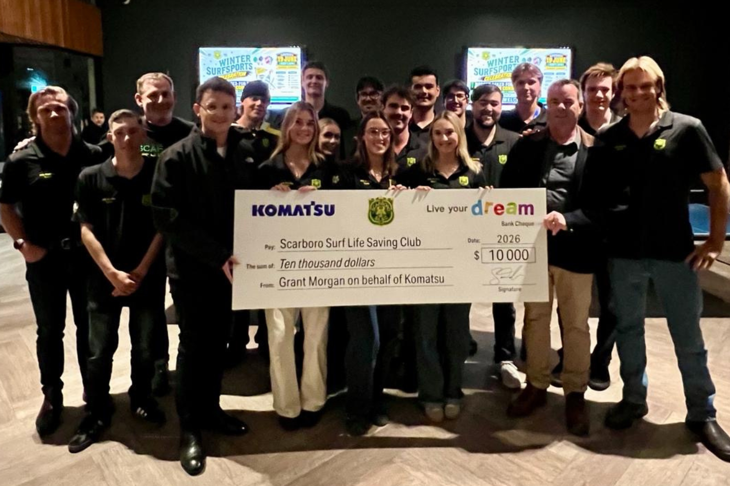 A group of people is gathered indoors holding a large check. The check is from Komatsu, indicating a donation of ten thousand dollars to the Scarboro Surf Life Saving Club. They are standing in a room with a patterned ceiling and posters in the background.