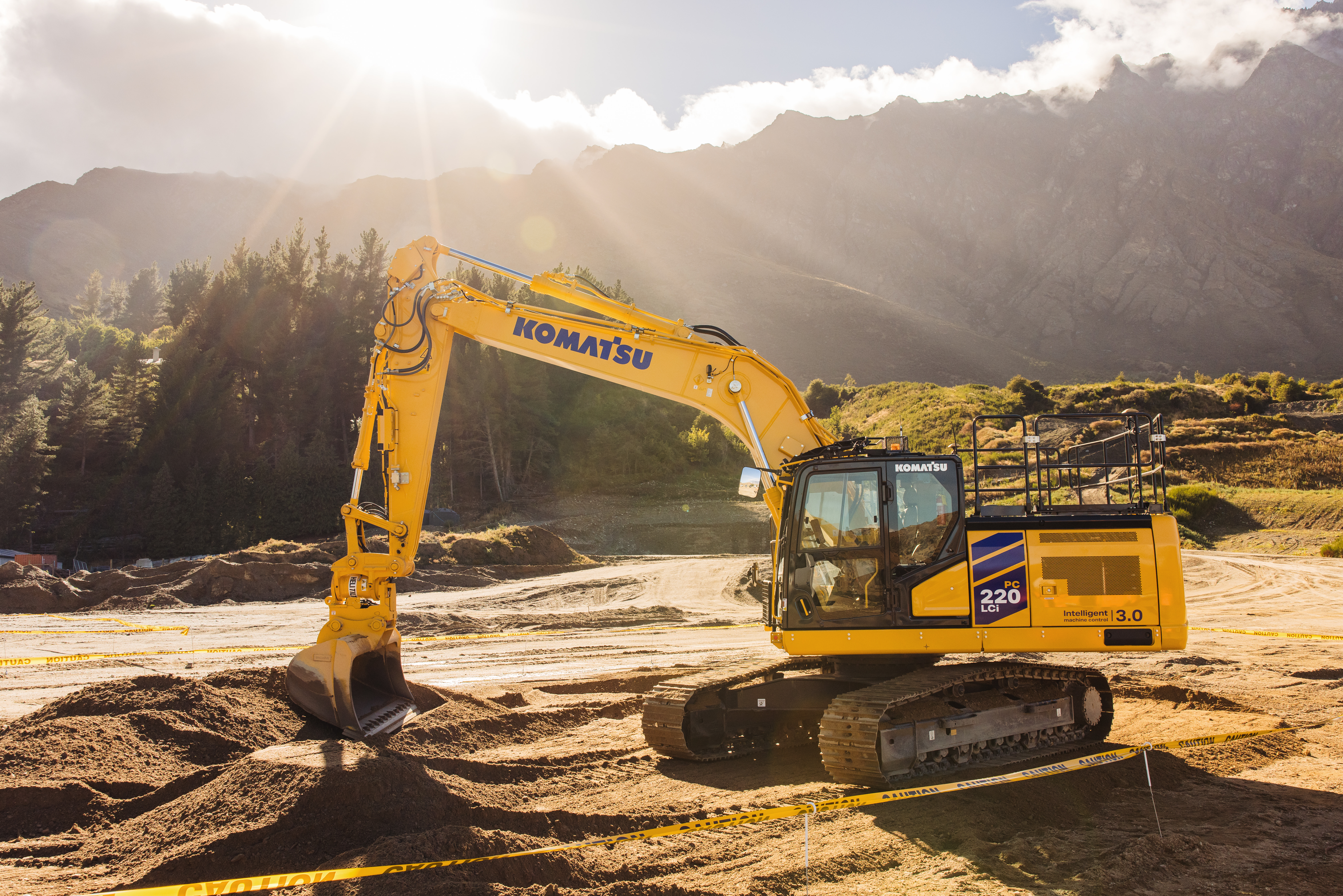 A Komatsu PC220LCi-12 excavator on a construction site on a sunny day. Mountains are visible in the background.