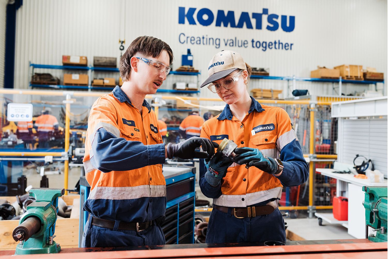 Two individuals in orange high visibility safety clothing are analysing a heavy machinery part in a workshop setting. The Komatsu logo, along with "Creating Value Together" are visible on the wall behind them.