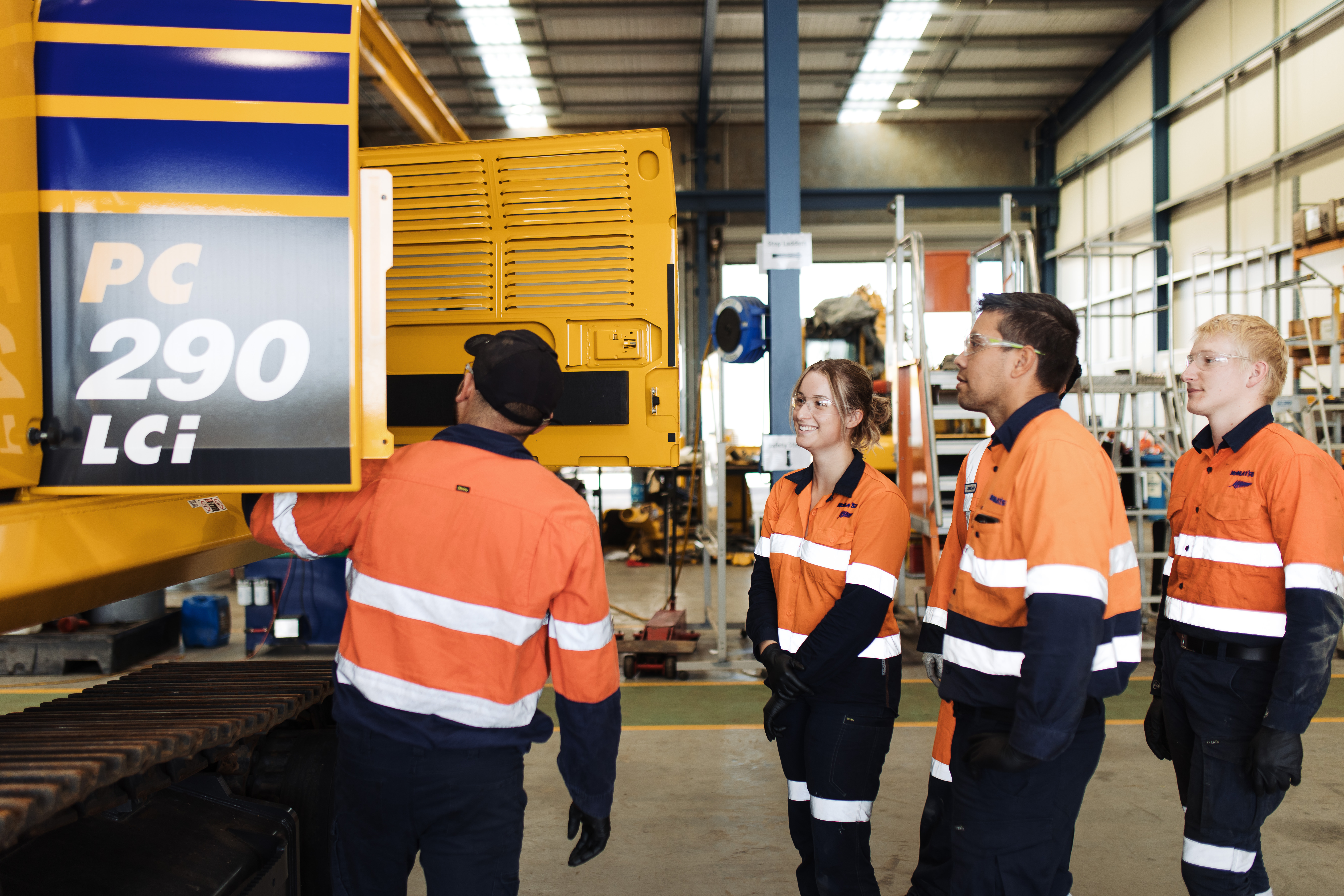 A group of Komatsu apprentices in orange high visibility workwear stand near a Komatsu PC290LCi machine in a workshop setting.
