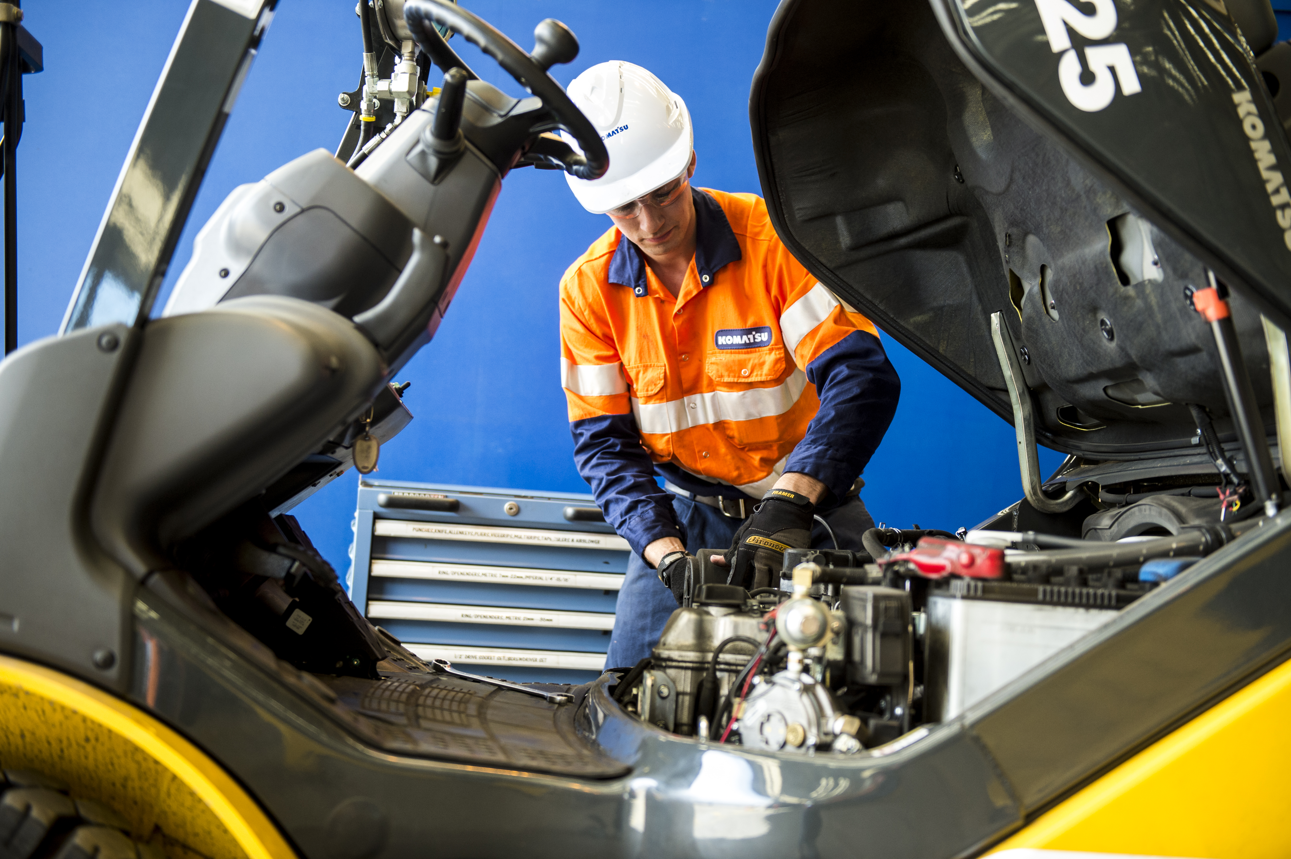Technician in a high-visibility orange uniform and white hard hat performs maintenance on a forklift engine, working inside the open compartment in a workshop setting.