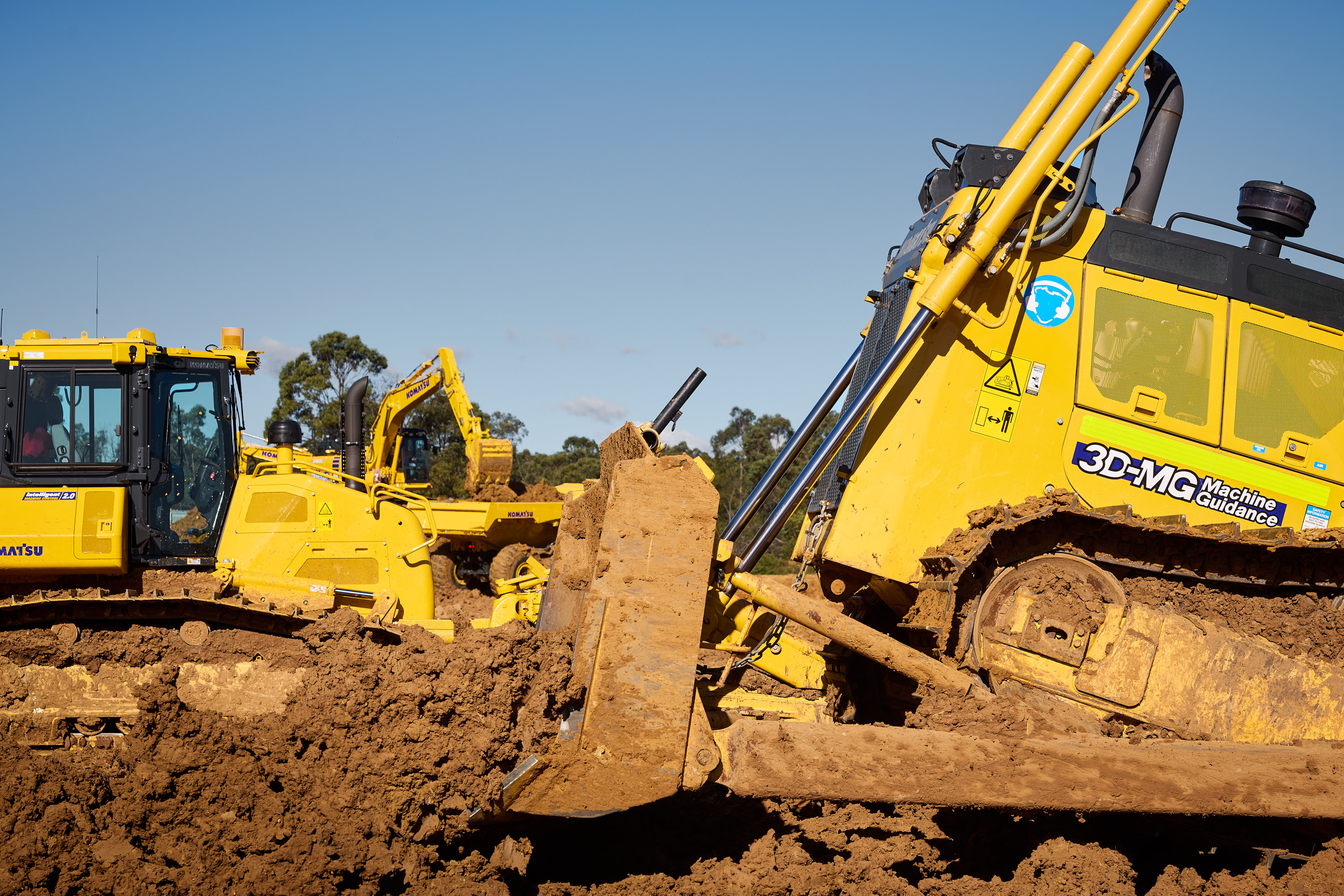 Two large yellow Komatsu bulldozers are actively moving soil at a construction site. The clear sky acts as a backdrop, while the focus is on the powerful machinery in motion.