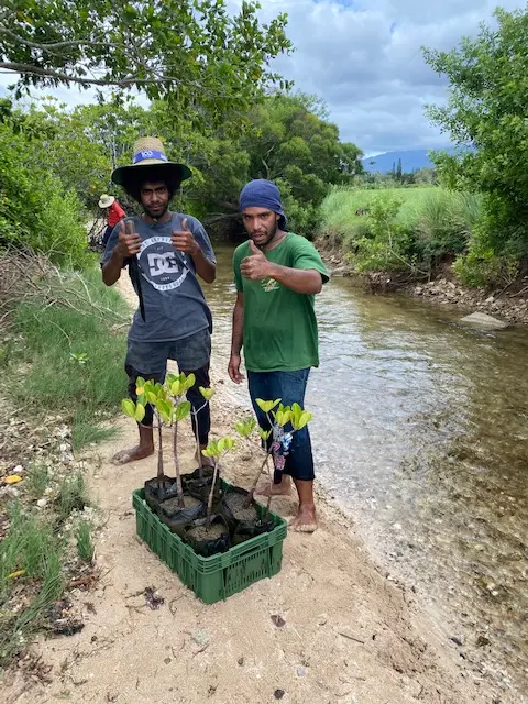 Two men giving thumbs-up at the side of a river with trees at their feet
