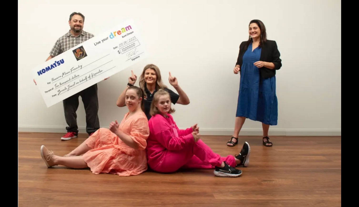 Four people sitting on a wooden floor and two standing behind them, with one person holding a large ceremonial check