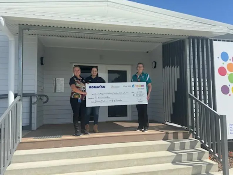 Three individuals standing in front of a building entrance, holding an oversized ceremonial check from Komatsu.