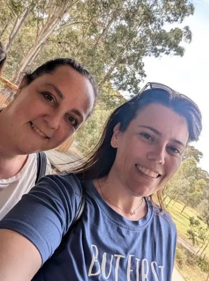 Two individuals posing for a selfie outdoors, with trees in the background.