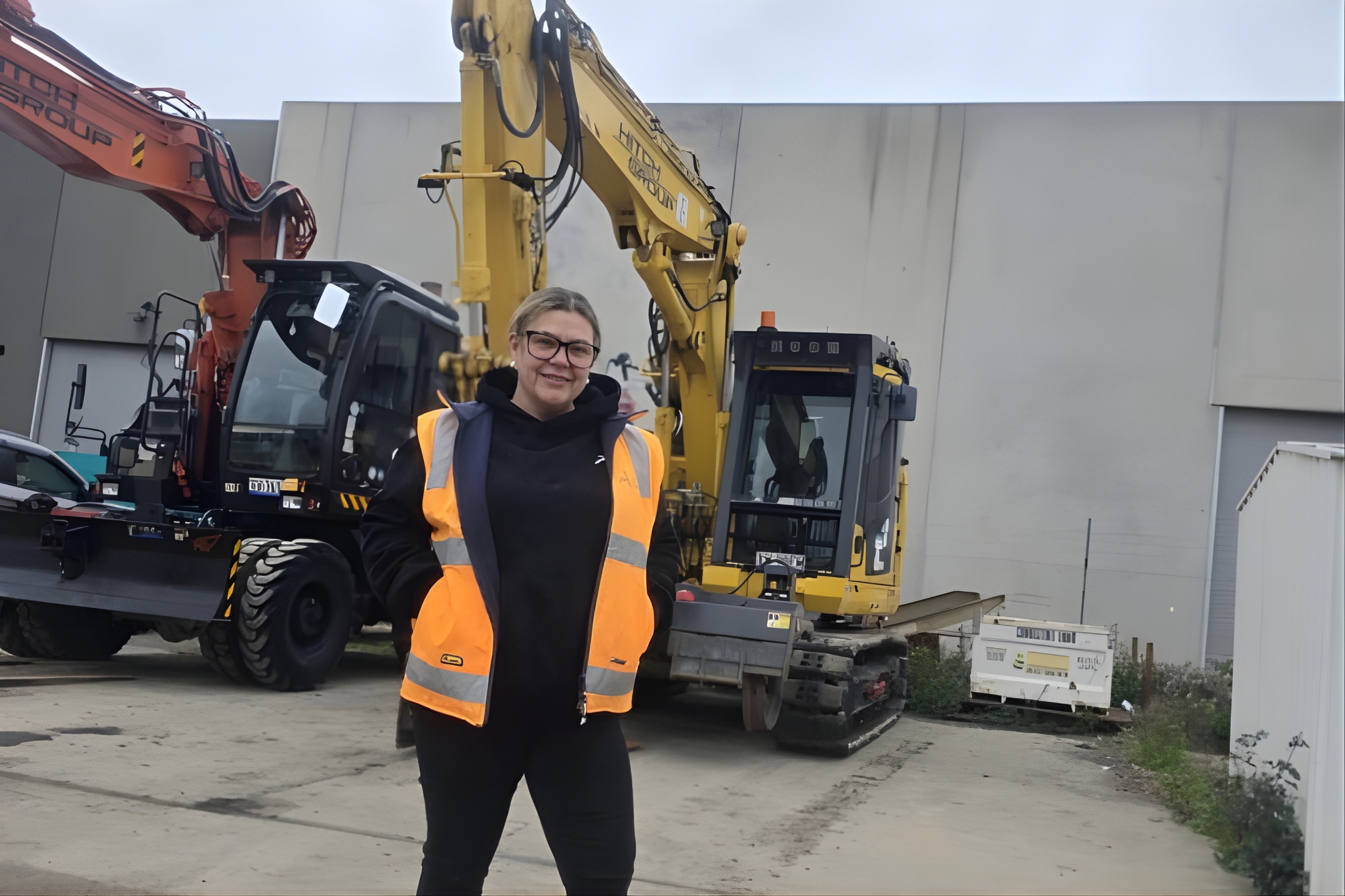 A person in a high-visibility vest and safety boots smiles in front of a yellow Komatsu excavator.