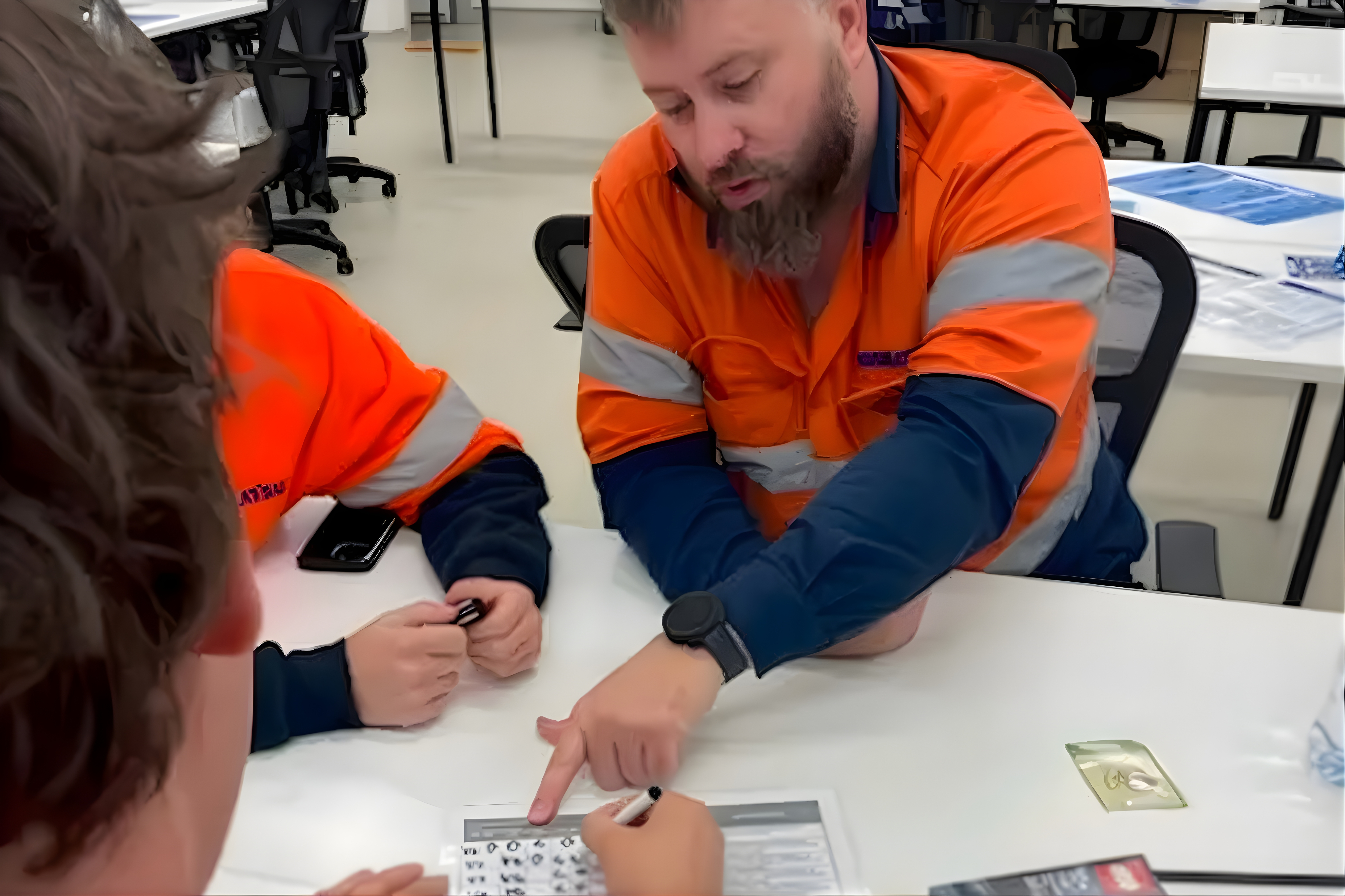 Two individuals in hi-vis vests are seated at a table, engaging in a discussion over a document with diagrams, in a classroom setting.