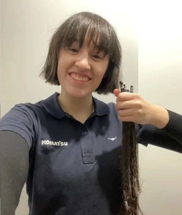 Person in a Komatsu polo shirt holding a long cut braid of hair and smiling at the camera.