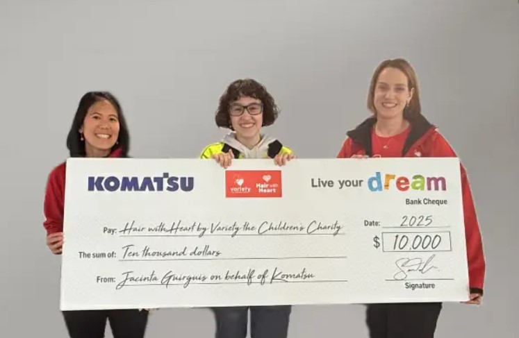 Three individuals hold a large ceremonial check from Komatsu to Live Your Dream for $10,000 addressed to Hair with Heart by Variety the Children's Charity.