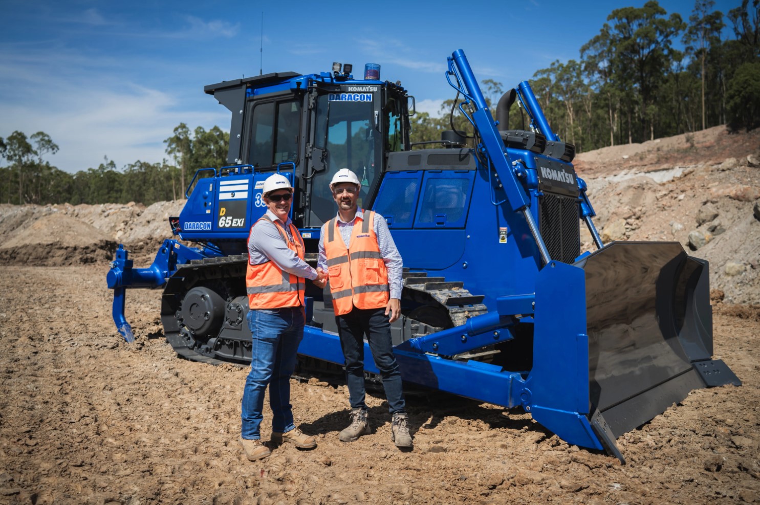 Two workers in safety vests and helmets stand in front of a Komatsu bulldozer at a construction site.