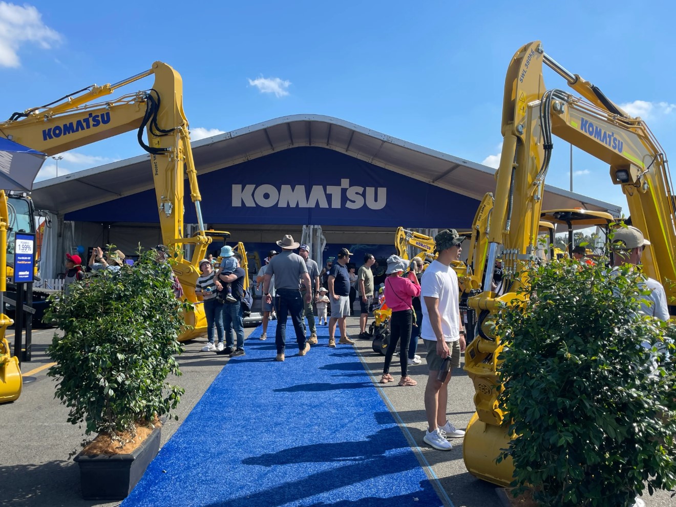 People gathered at a Komatsu equipment exhibition with multiple yellow excavators displayed under a large tent bearing the Komatsu logo.