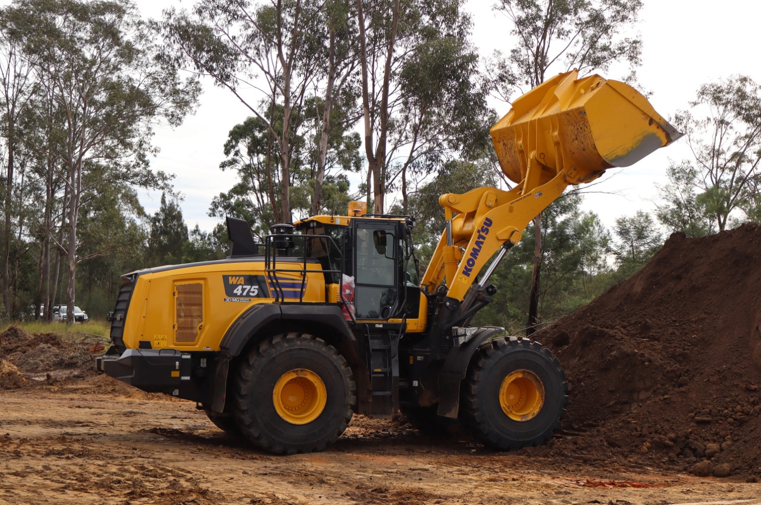 Komatsu WA475 wheel loader dumping soil in a wooded area.