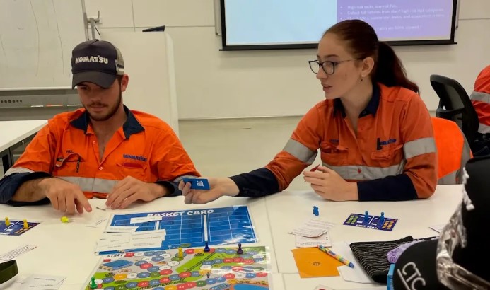 Two individuals in high-visibility orange workwear are seated at a table playing a board game.