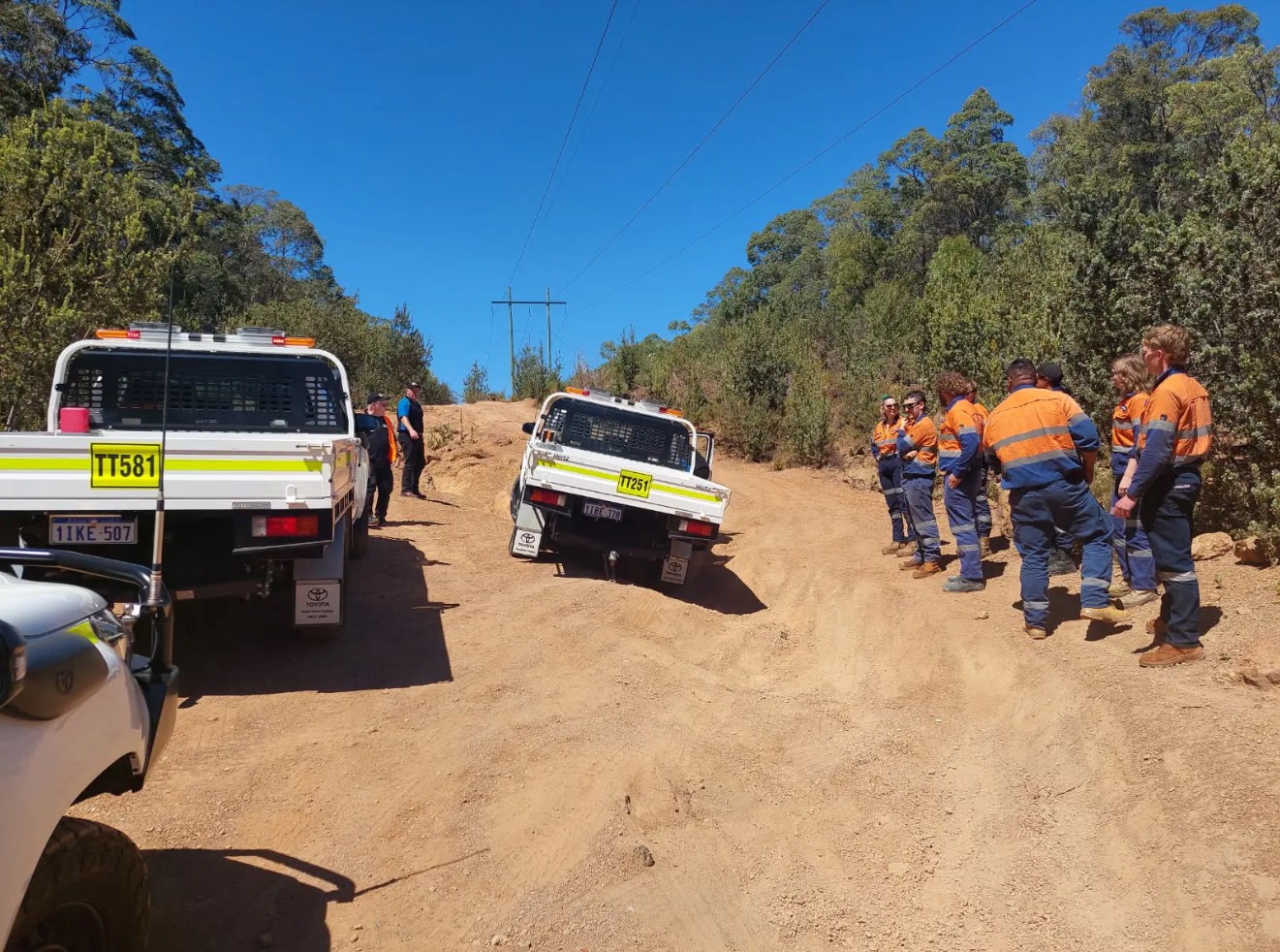 Individuals in high visibility workwear standing next to vehicles at a rural roadside scene, on a dirt road under a clear blue sky.