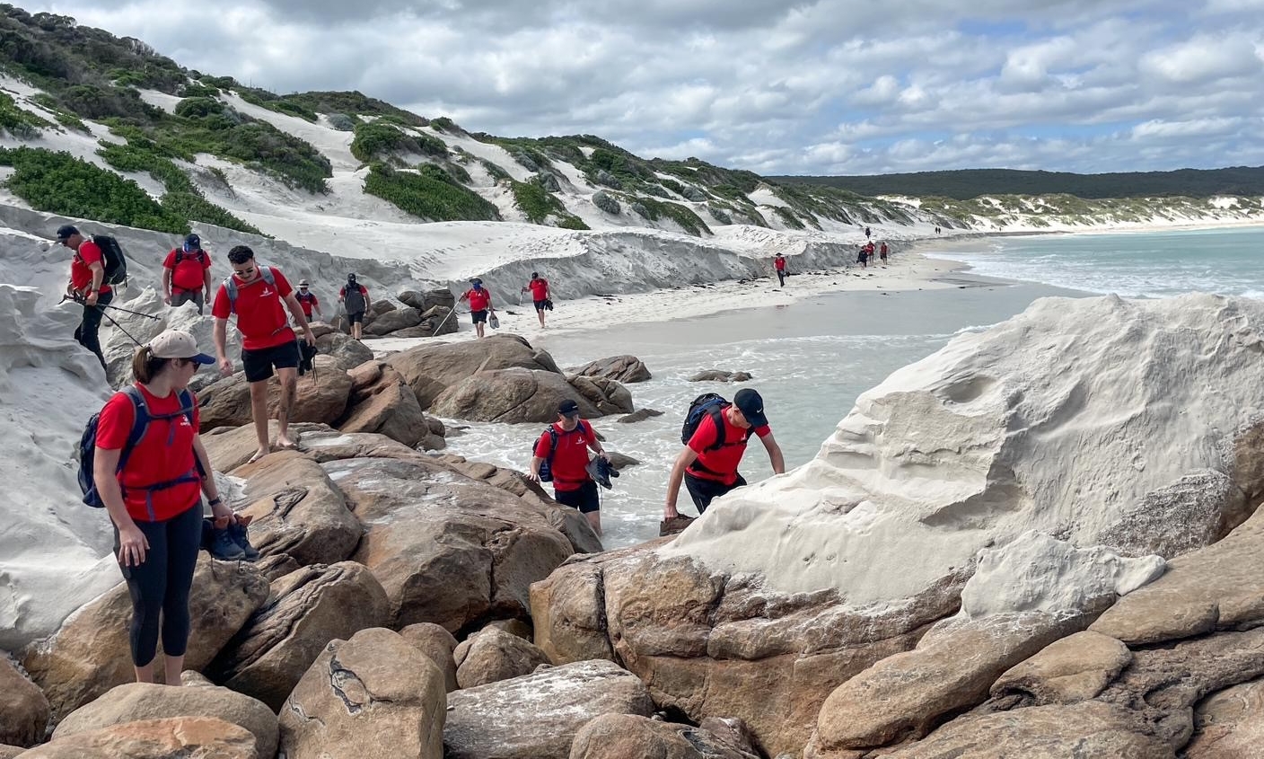 A group of people wearing red shirts exploring a rocky coastal area with white sand dunes under a cloudy sky.