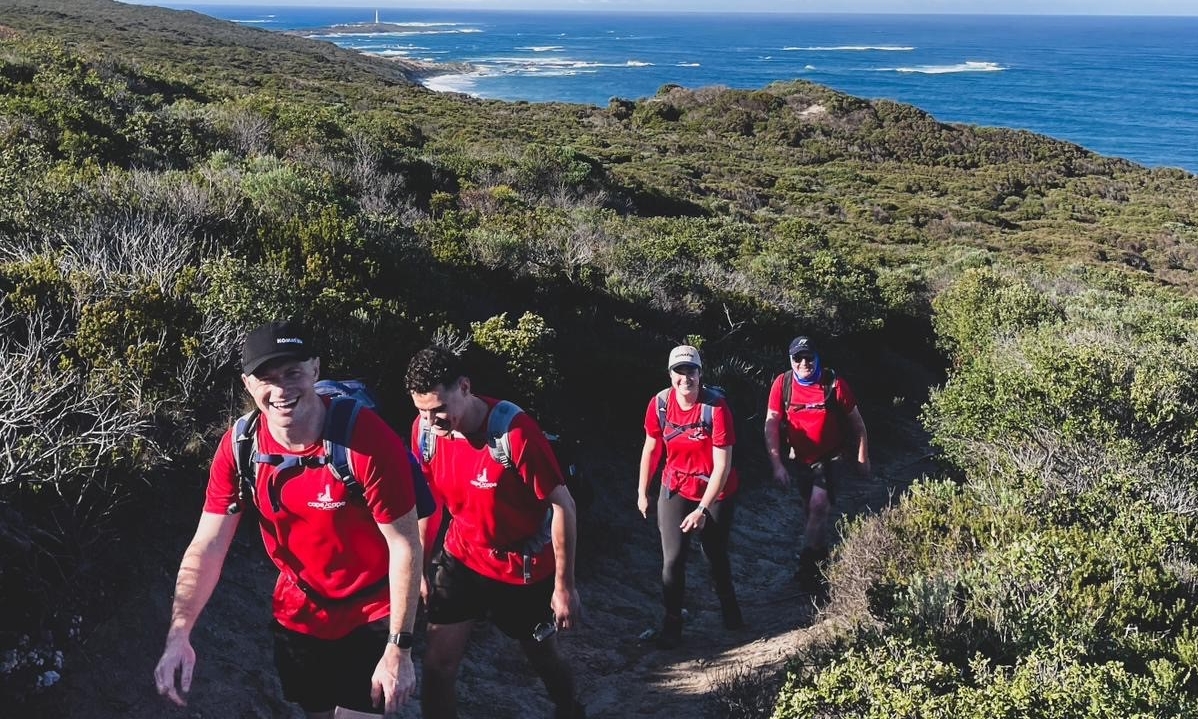 Four individuals wearing red shirts hiking on a coastal trail with lush greenery and a view of the ocean in the background.