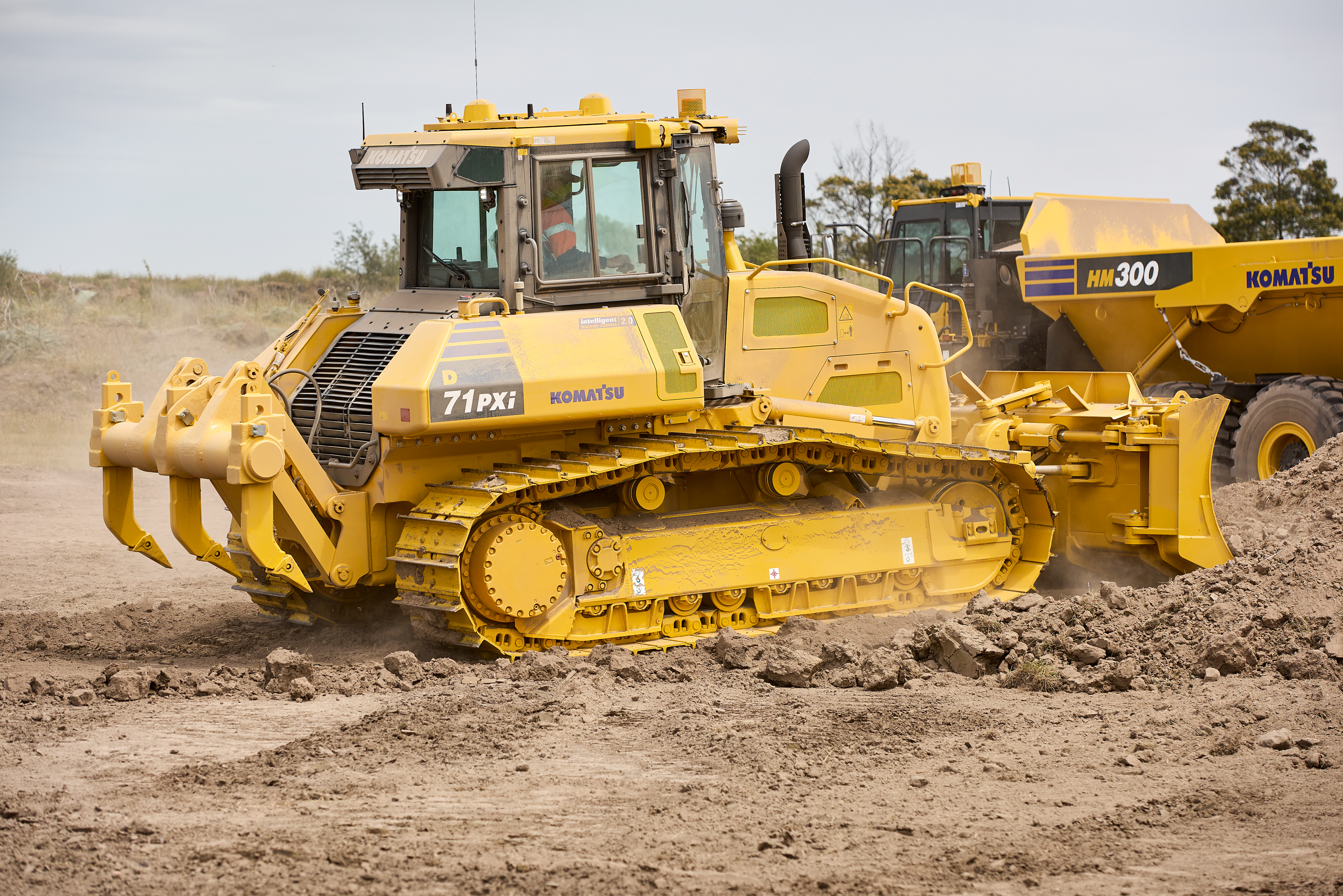 A Komatsu D71PXi bulldozer operates on a dirt field alongside a Komatsu HM300 articulated dump truck.