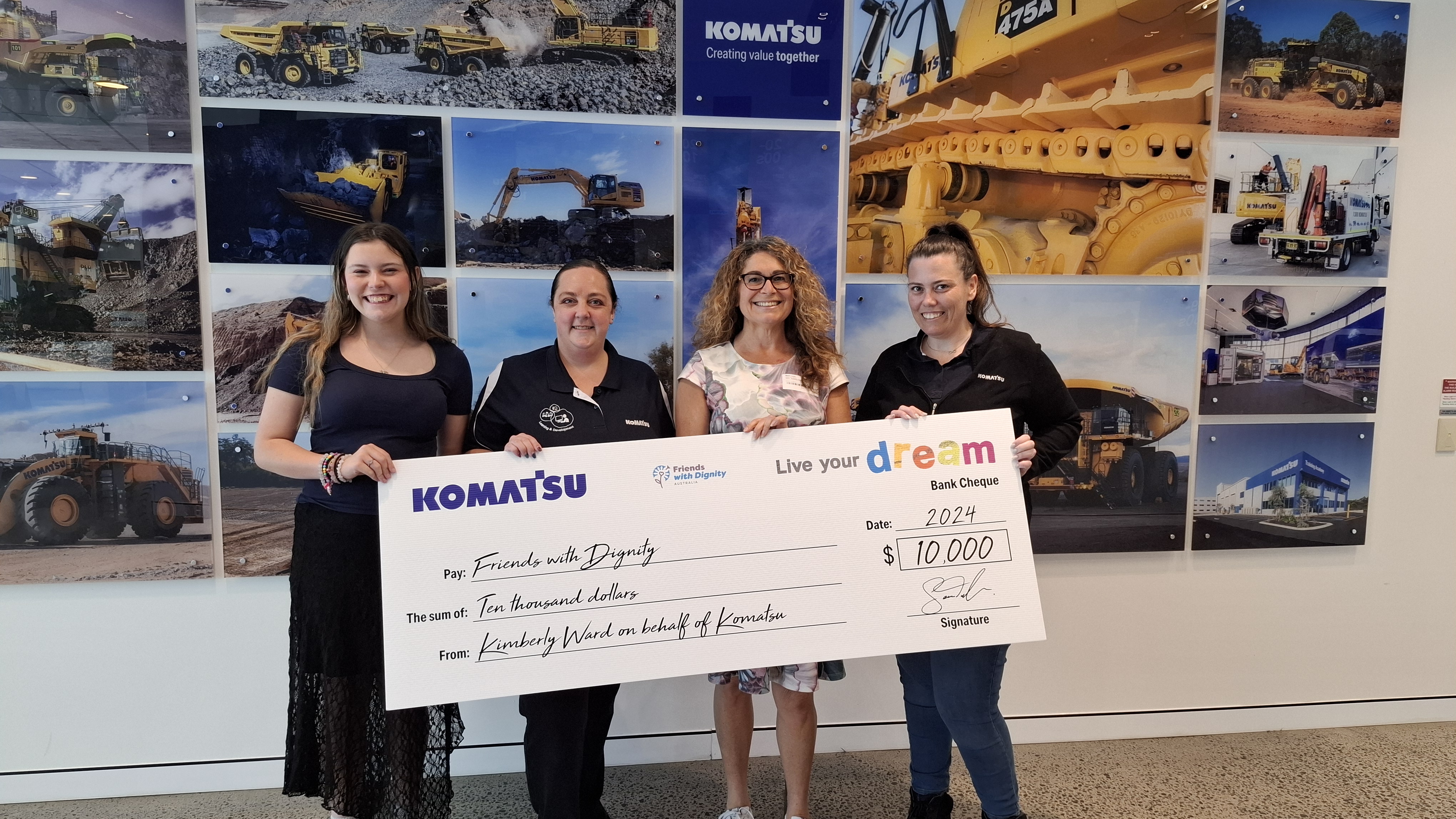 Four people posing in an office with large photo banners of construction equipment in the background. They are holding a large promotional check from Komatsu for $10,000, dated 2024. The check is made out to "Friends with Dignity.