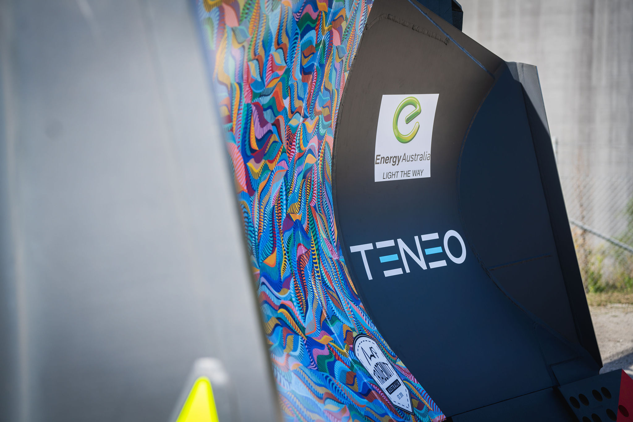 A partial view of a bulldozer blade featuring colorful abstract patterns with branding logos of EnergyAustralia and Teneo displayed prominently.