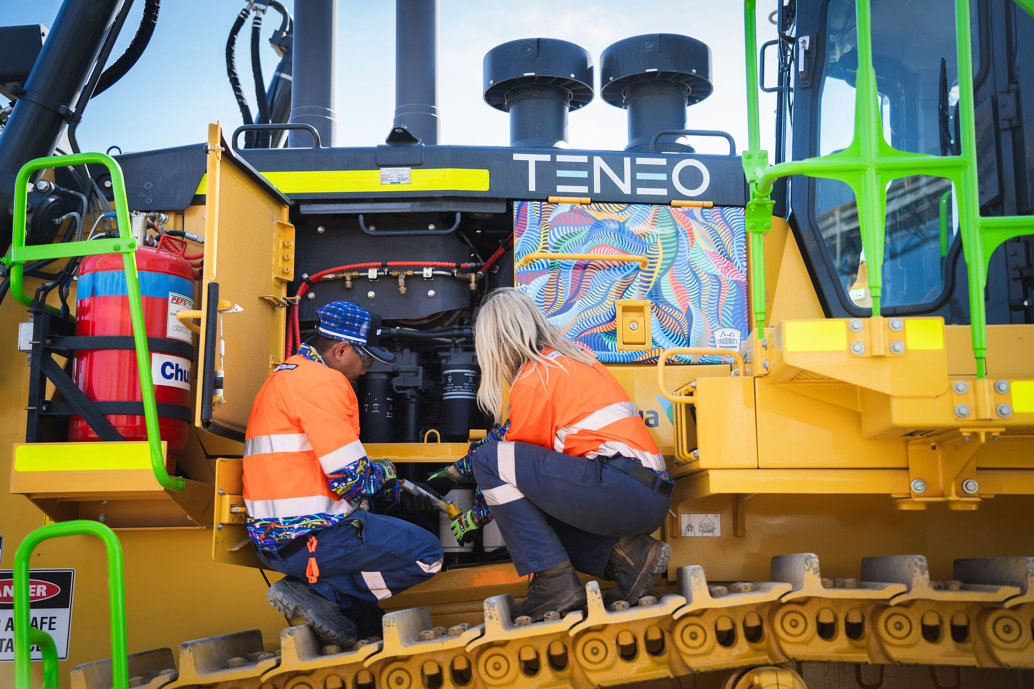 Two workers in high visibility clothing examine the engine of a Teneo-branded construction vehicle.