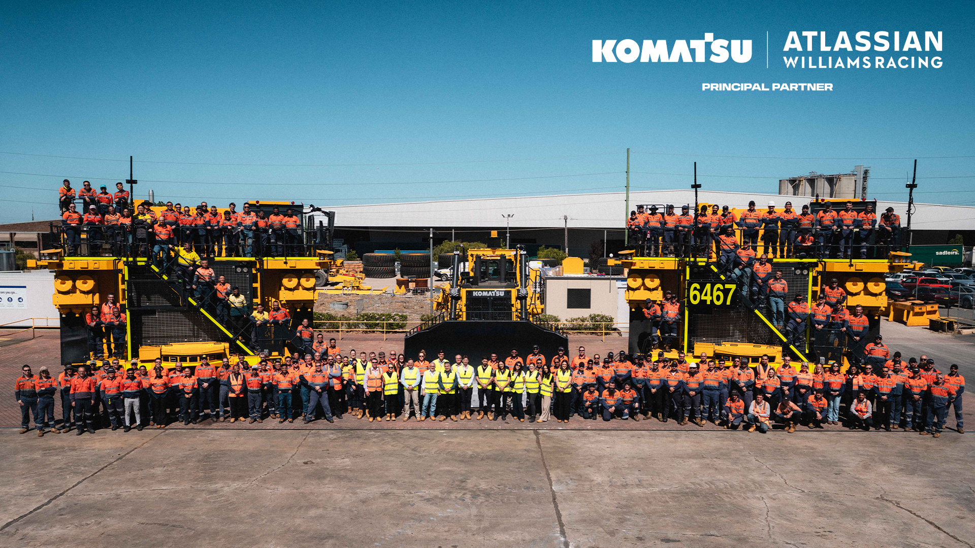 Group of workers in orange and yellow safety vests gathered in front of large yellow Komatsu machinery, with "Komatsu" and "Atlassian Williams Racing" logos visible.