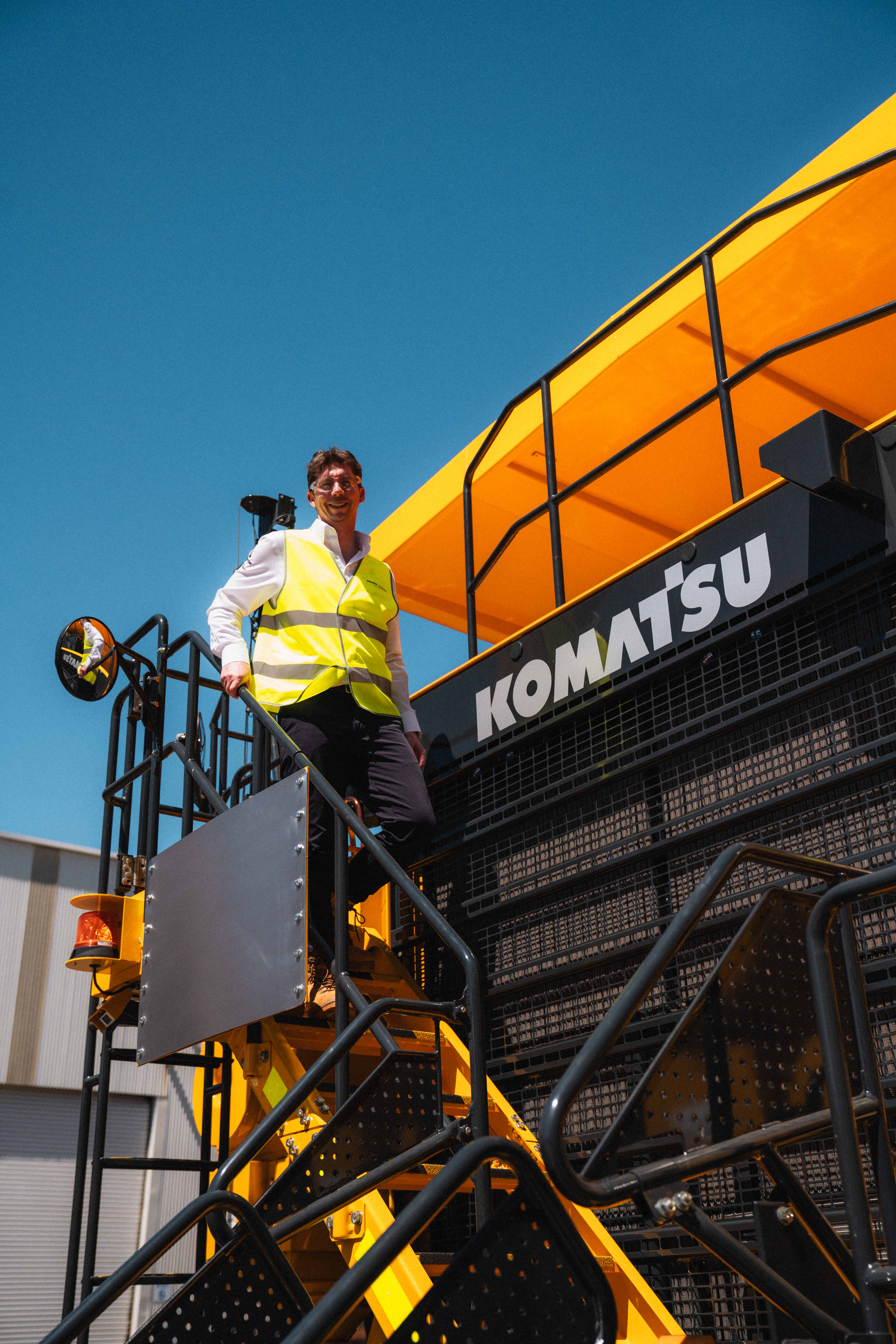 A person in a high visibility vest standing on a Komatsu industrial machine under bright sunlight.