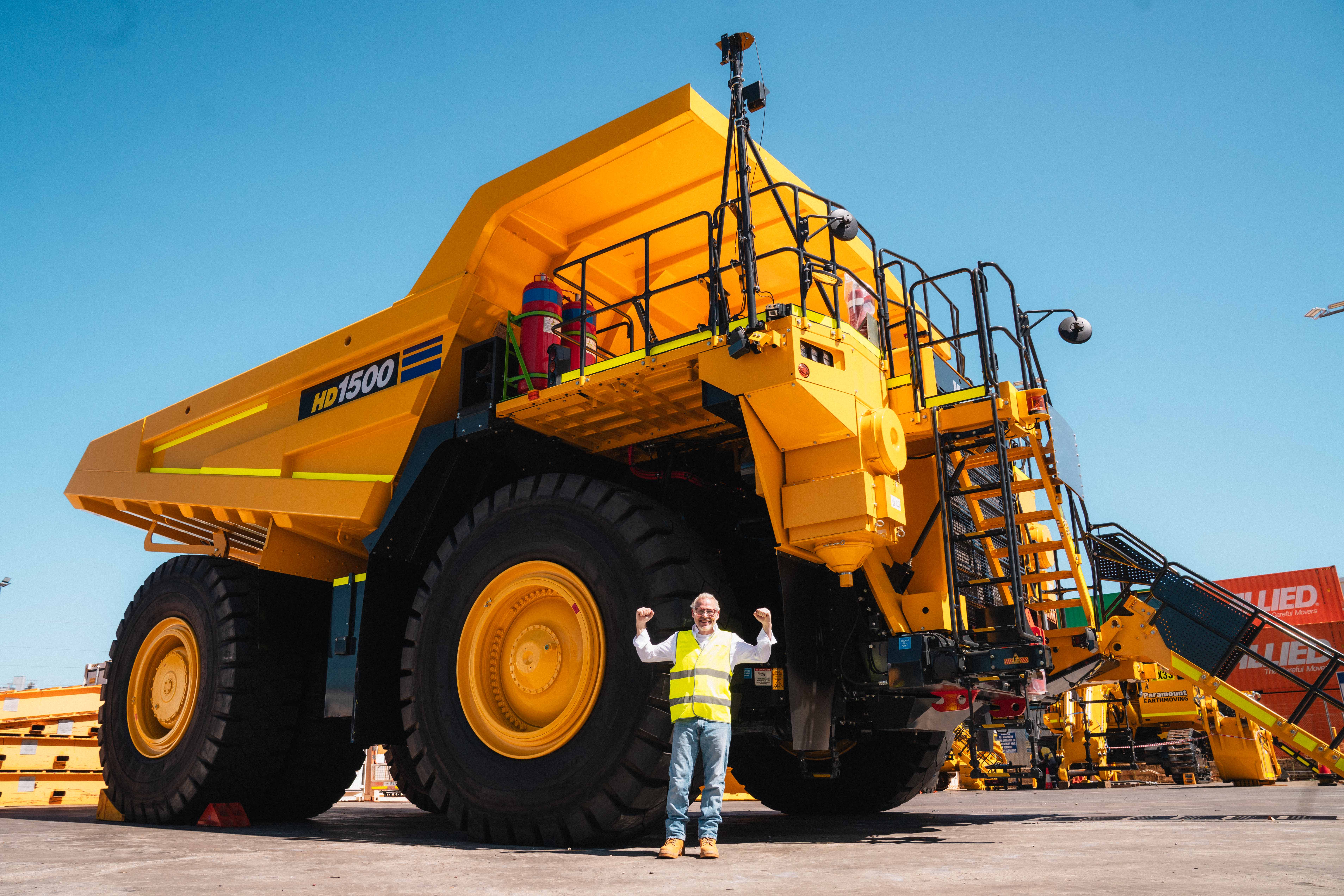 Worker in safety vest and helmet stands smiling in front of a large Komatsu truck on a sunny day at an industrial site.