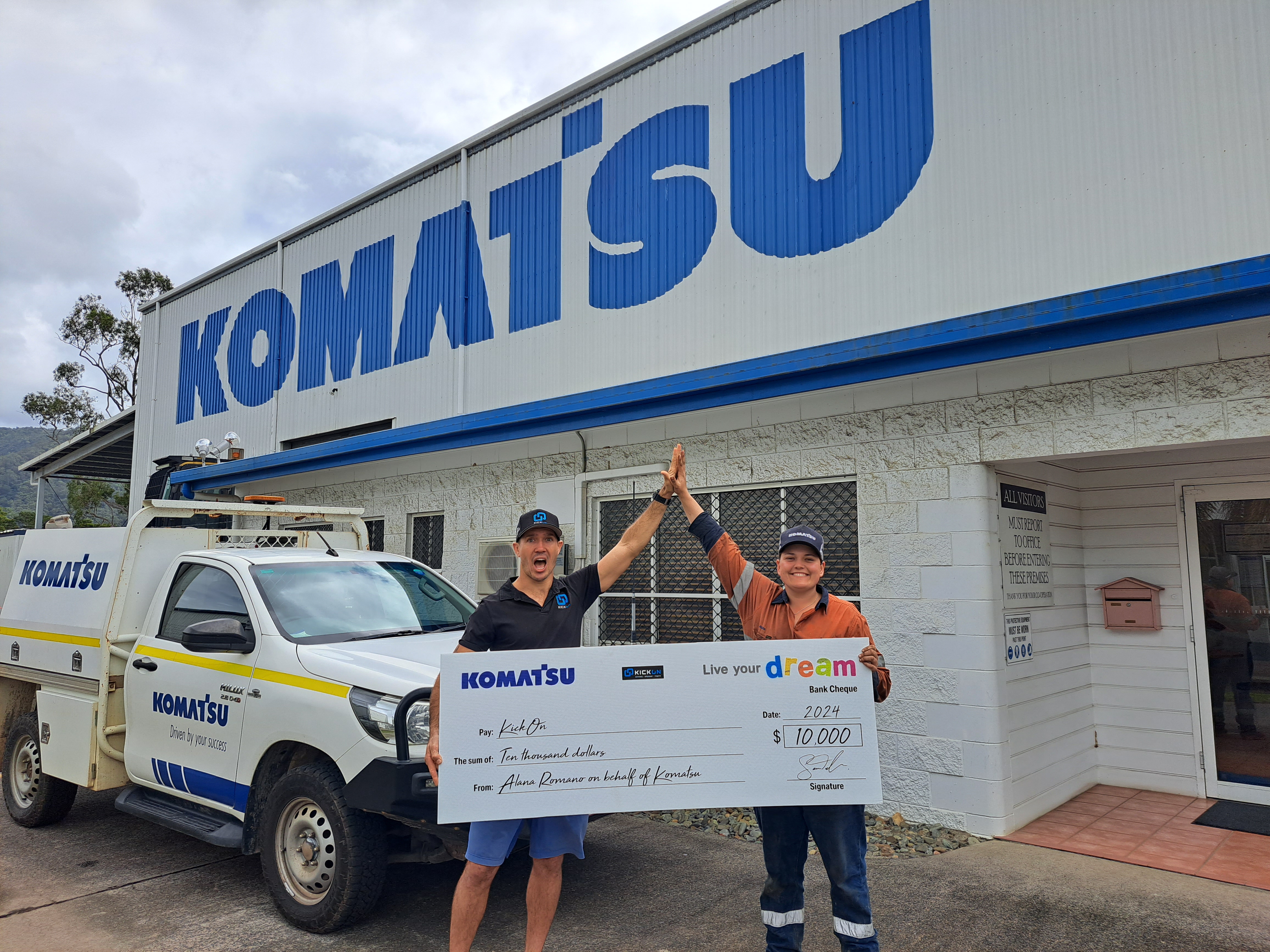 Two individuals celebrating with a handshake in front of a Komatsu building, holding a large check for $10,000 marked "Live Your Dream".
