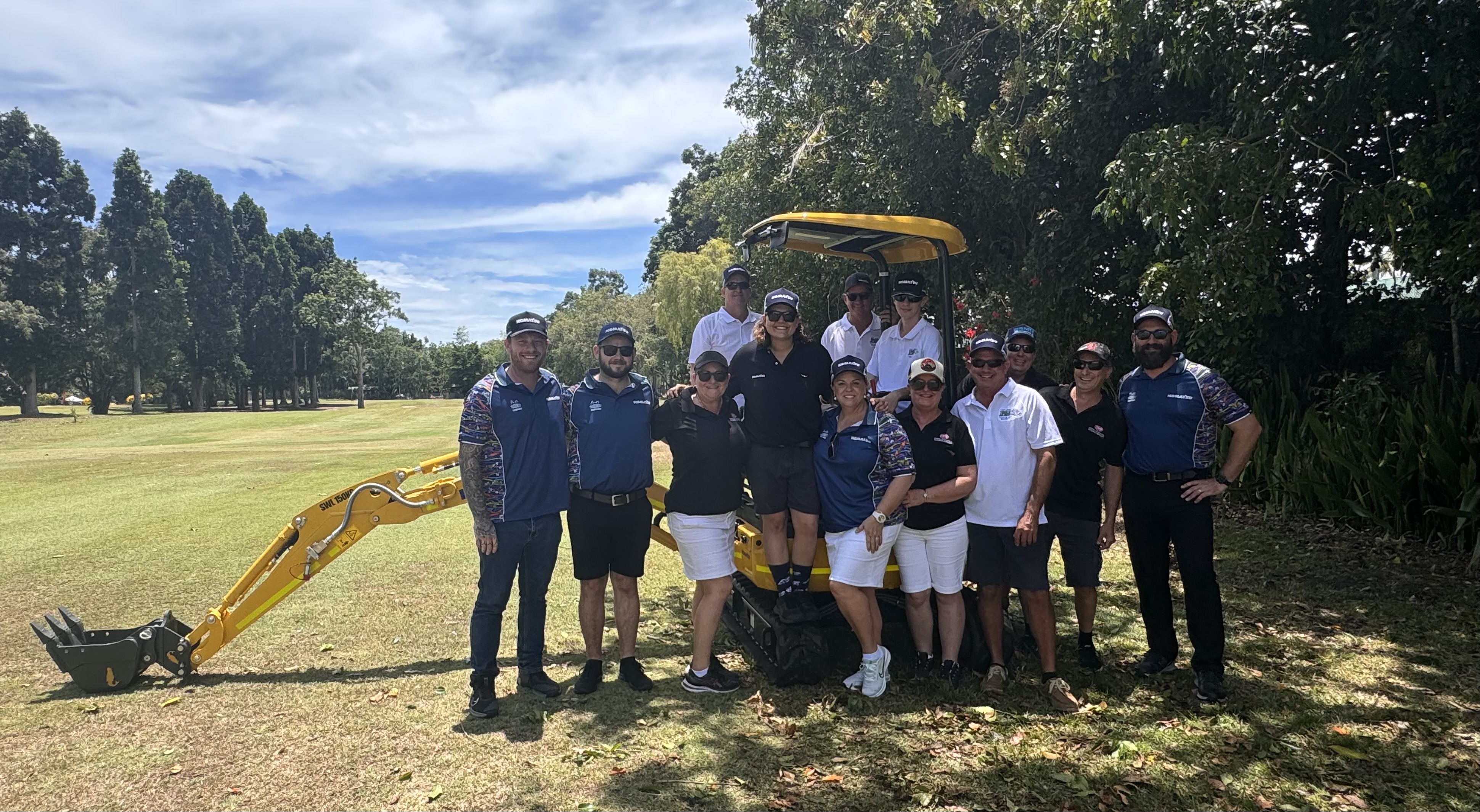 Group of people in uniform standing in front of a yellow excavator in a park.