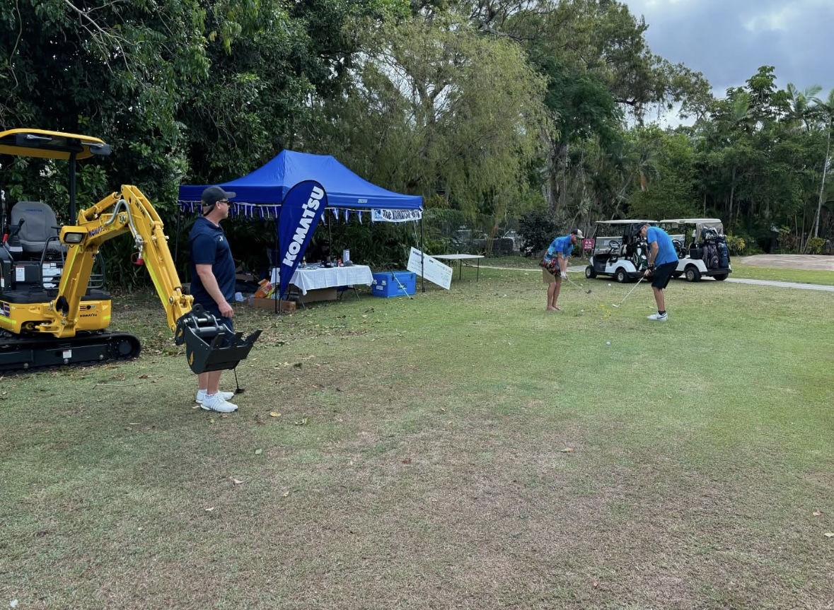 An outdoor event with three people and a Komatsu excavator at a demonstration area under a branded tent. One person carries a bucket near the excavator, while the other two are tossing a ball in a grassy field.