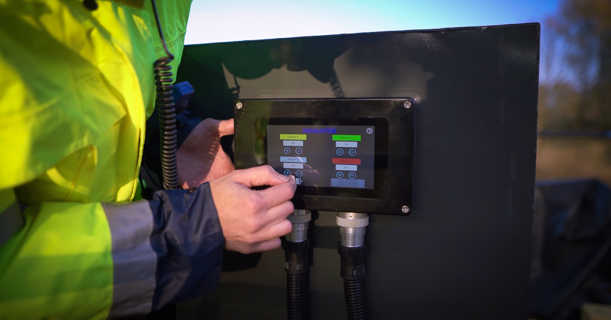 Person in a high‑visibility jacket operating an outdoor control panel with a touchscreen display.