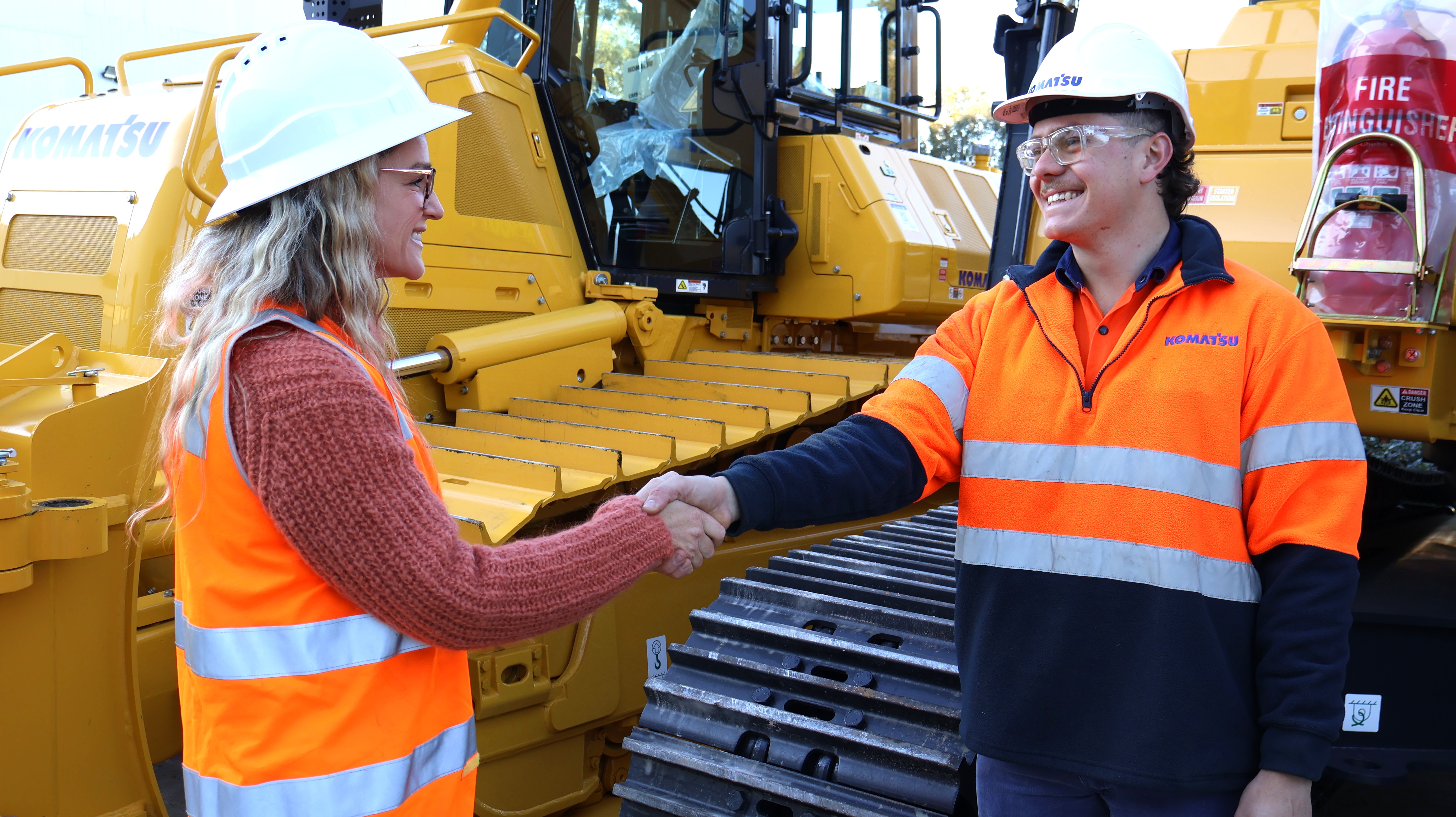A Komatsu technician shaking hands with a yellow vest man in front of Komatsu equipment
